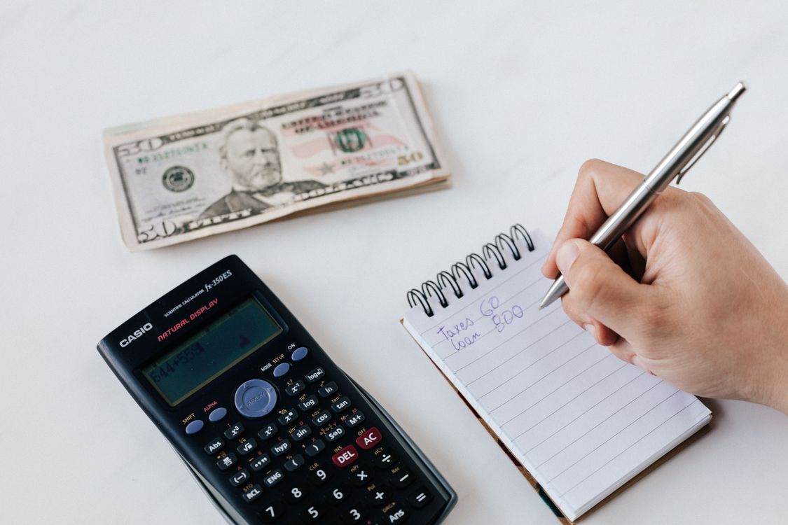 Pessoa anotando valores em um caderno ao lado de uma calculadora e cédulas de dólar.