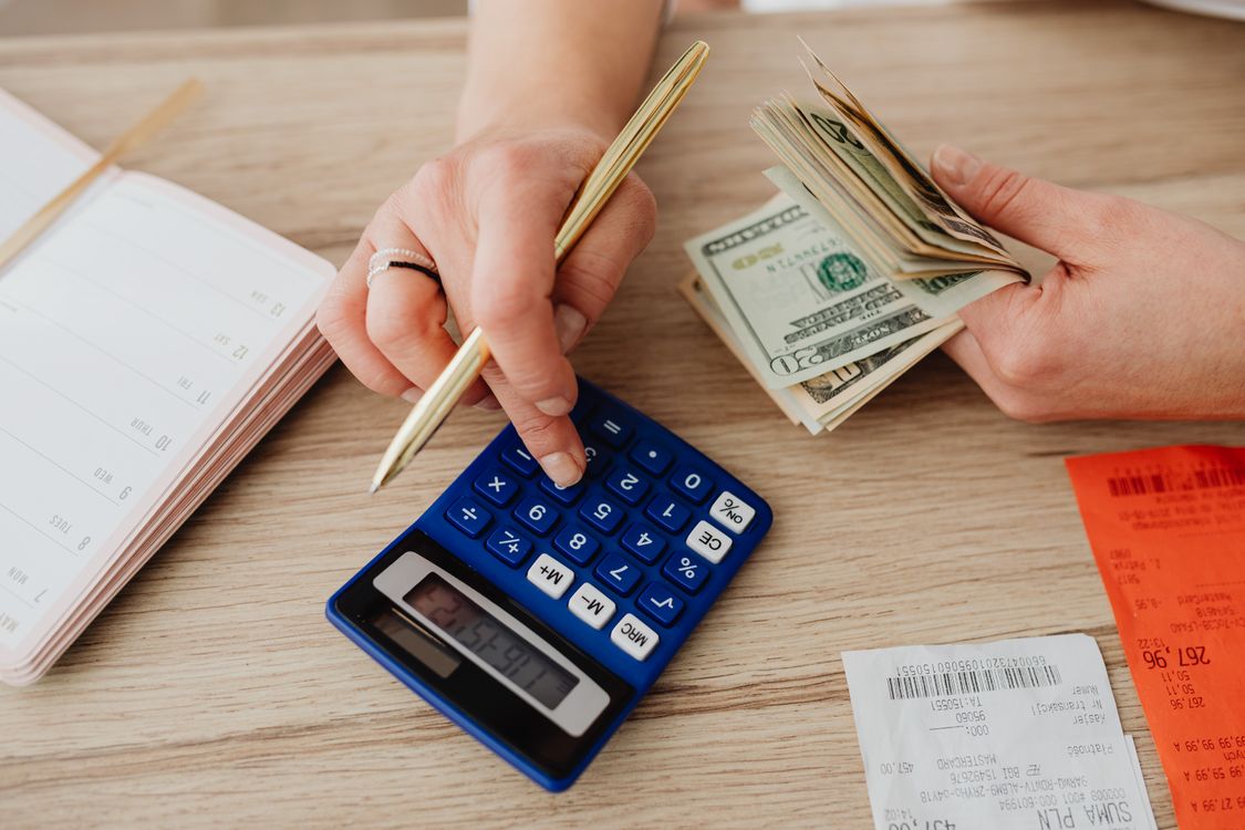 Mãos usando uma calculadora ao lado de recibos, agenda e cédulas de dólar, em uma cena ilustrativa de acompanhamento das contas.